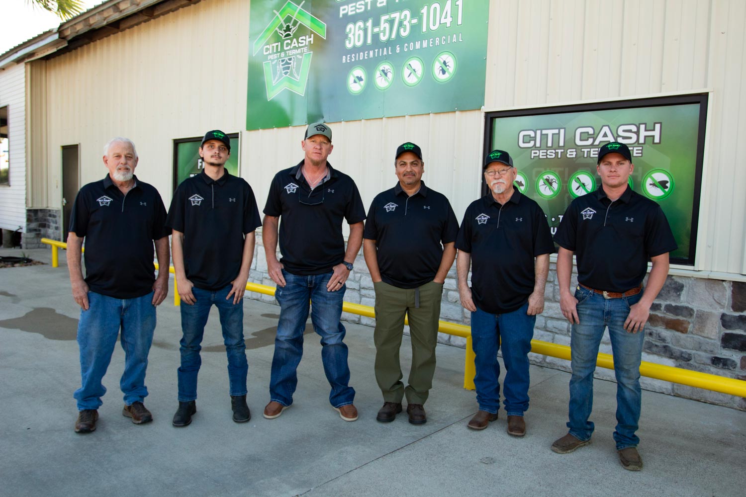 Six men in matching black shirts and hats stand in front of a building with pest control signs.