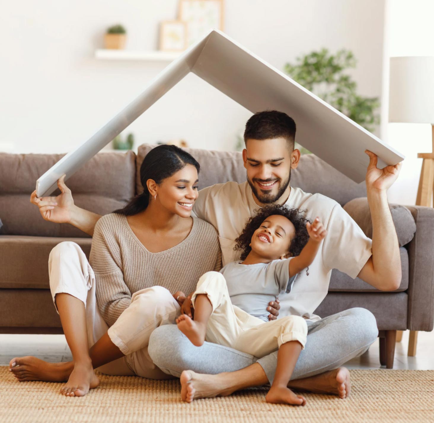 Smiling family of three sits on the floor, holding a roof-shaped panel over their heads in a cozy living room.