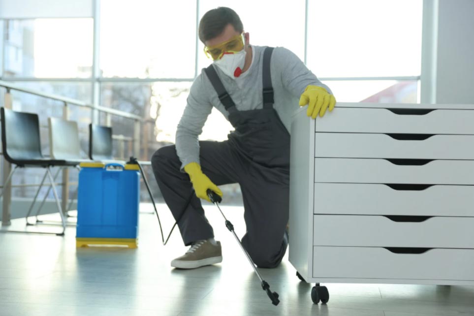 A person wearing gloves and a mask sprays disinfectant near a white cabinet in a bright room.