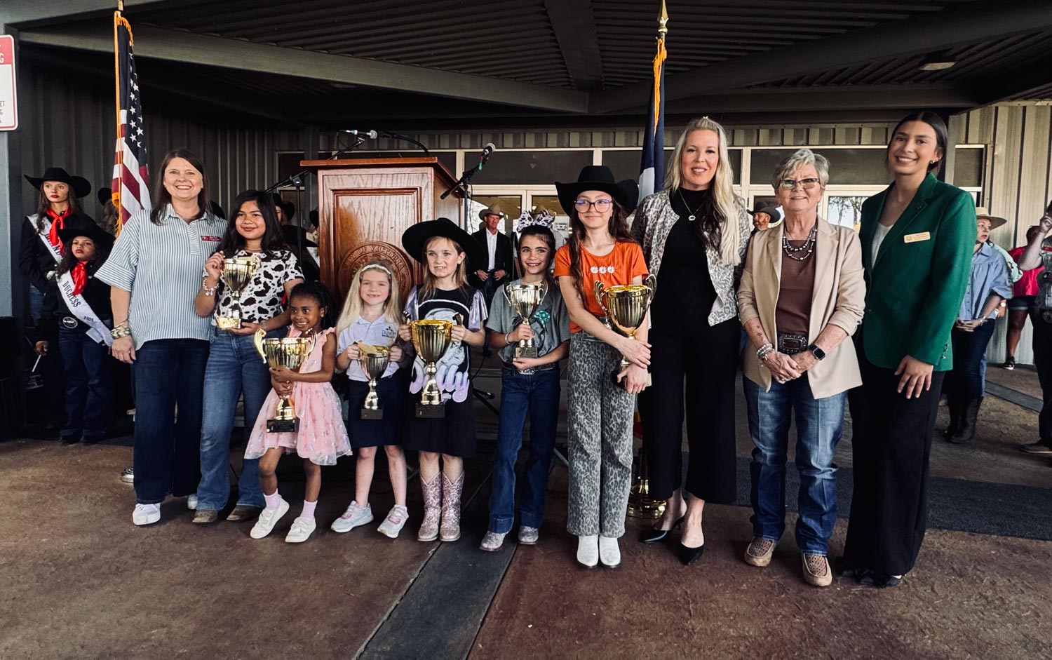 A group of children with trophies and adults stand together, smiling, at an indoor ceremony or event.