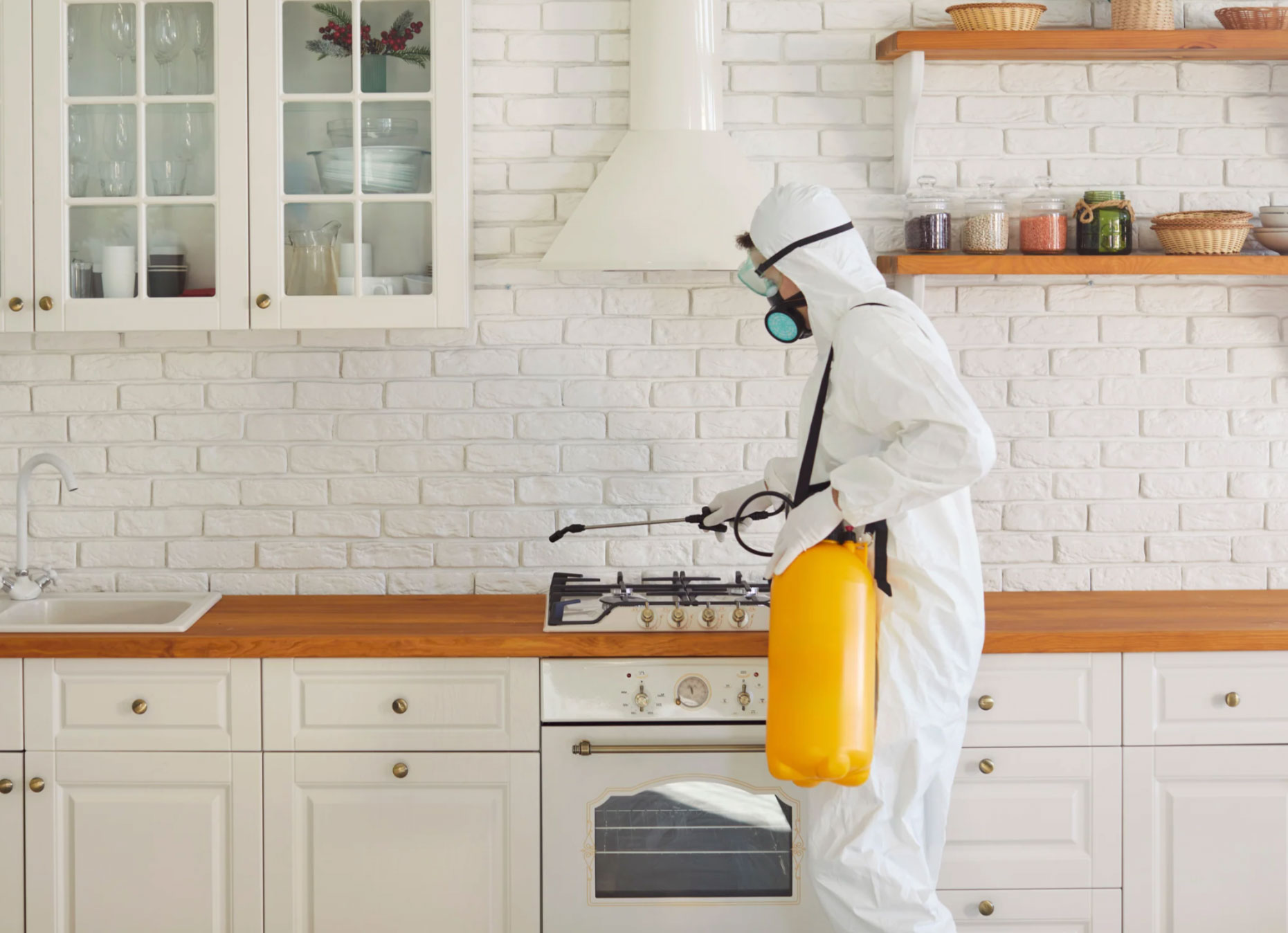 Person in protective suit spraying pesticide in a white kitchen with wooden countertops and open shelves.